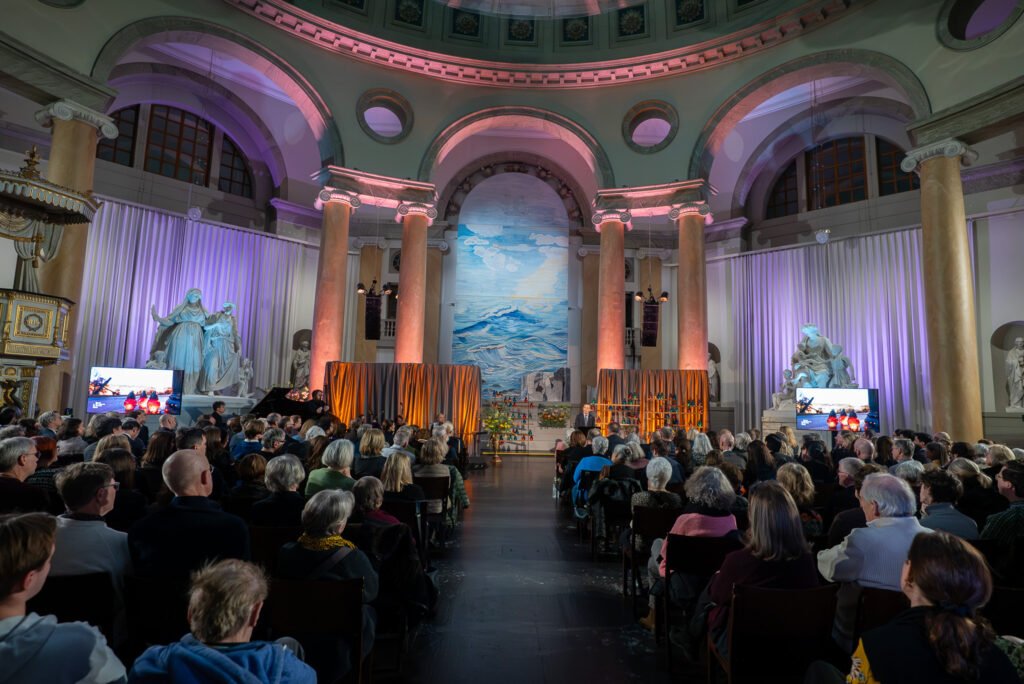 Wide view of a full audience in an ornate domed hall facing a stage lit in purple and orange
