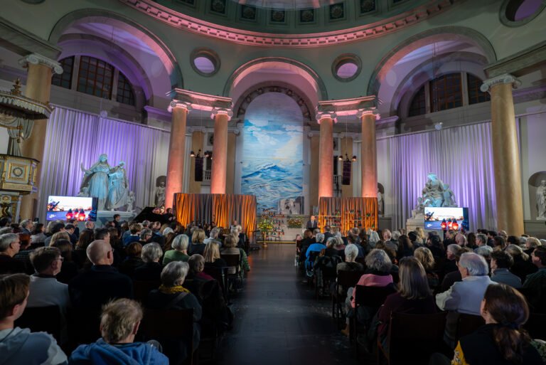 Wide view of a full audience in an ornate domed hall facing a stage lit in purple and orange
