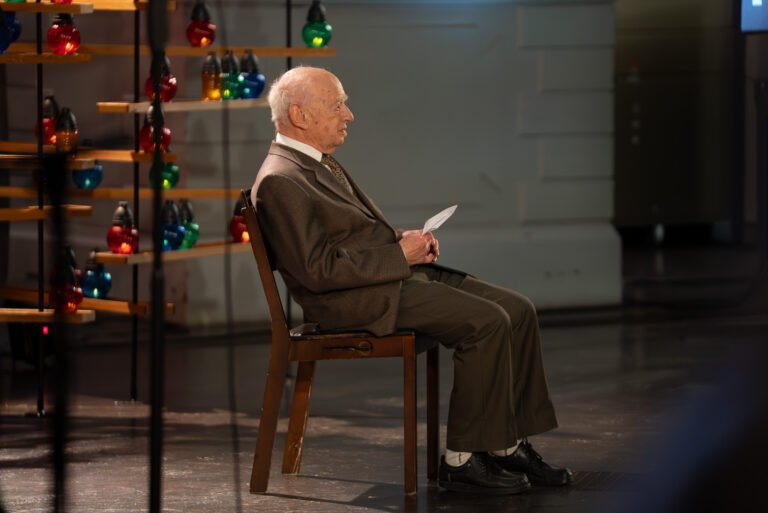 Older man in a brown suit seated on a wooden chair on stage, holding a card, beside shelves of coloured glass lanterns