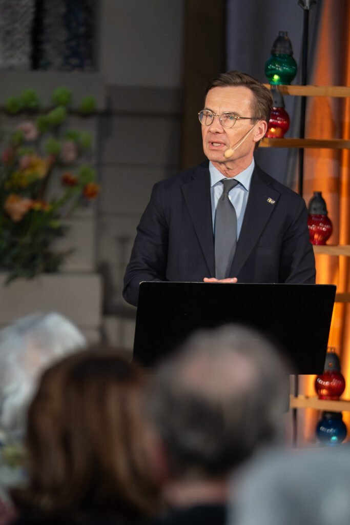 Speaker in a dark suit addressing an audience from a podium, with warm stage lighting and flowers behind him