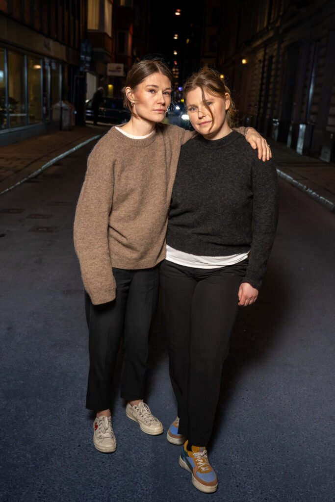 Two women in knitted jumpers standing arm in arm in the middle of a quiet Stockholm street at night